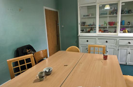A room with a table in the middle and six chairs around it. At the back of the room there is a cabinet with shelves behind glass doors, and drawers and cupboards.