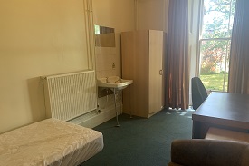 A room with a single bed, a sink with a mirror over it, and a wardrobe against one wall. There is a desk against the opposite wall with an office chair and an armchair near it.