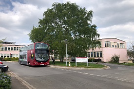 A red and grey bus parked on a road near a tree. There is a large pink two-storey building behind the tree and a sign nearby saying 'Churchill Building'.