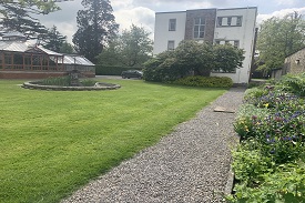 A path leading up the side of a lawn towards a white three-storey building. There is a fountain on the lawn and a greenhouse on the other side of it.