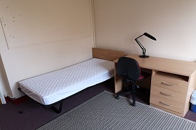 A single bed to the left of the room. Next to the bed is a desk with a lamp on it, and a chair.