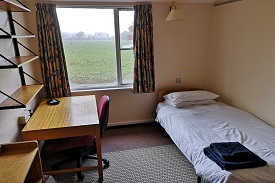 A single bed to the right of the room with a desk, chair and shelves the other side of the room, a window with curtains with a view of gardens.