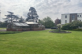 A fountain on a lawn with a white three-storey building behind it. There is a greenhouse to the side of the lawn.