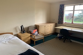 A room with a single bed in one corner and a desk against the opposite wall under a window. Against the wall between them there is a bedside table, an armchair, and a small cabinet with cupboards and drawers.