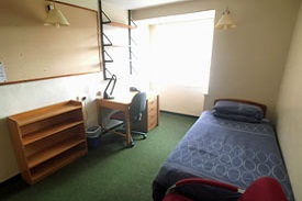 A room with a single bed in one corner, with an armchair at the foot of the bed. There is a desk against the opposite wall, with an office chair, a set of 4 shelves on the wall above it, a set of drawers beneath it, and a small bookcase next to it.