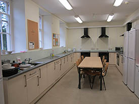 A large kitchen with three ovens and hobs, three sinks, three fridge freezers and lots of cupboards and drawers. There is a table in the centre of the room with eight chairs around it.
