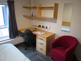 A room with a desk against one wall and a bed opposite. The desk has an office chair next to it, a set of shelves above it, and an armchair to the side.