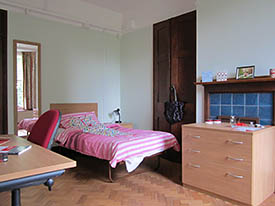 A room with a single bed and mirror against one wall, and a wardrobe, mantlepiece and chest of drawers on the adjacent wall. There is a desk and an office chair opposite.