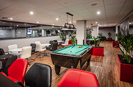A large room with lots of table surrounded by chairs, lots of potted plants, and two pool tables in the centre of the room.
