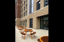 an exterior photograph of the outside of Metal Works A block, 5 orange picnic tables on concrete in front of a beige brickwork building with a number of windows and doors.