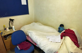 A single bed in the corner of a room with a bedside table and an office chair next to it.