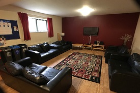A room with five black leather sofas arranged around a rug in the centre of the room, and a televion on the far wall.
