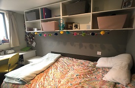 A room with a single bed and a set of shelves on the wall above it. At the foot of the bed there is a desk with an office chair.