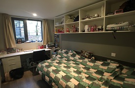 A room with a single bed and a set of shelves on the wall above it. At the foot of the bed there is a desk with an office chair and some drawers beneath it.