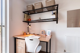 A desk in the corner of a room with an office chair next to it, a set of drawers below it and two shelves on the wall above it.
