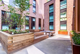 A paved courtyard with a raised wooden platform and seating, a ping pong table, and several plants in wooden holders. The courtyard is enclosed by two multi-storey red brick buildings.