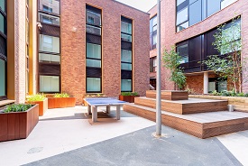 A paved courtyard with a raised wooden platform and seating, a ping pong table, and several plants in wooden holders. The courtyard is enclosed by a three-storey red brick building.