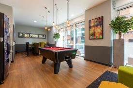A room with a vending machine, a pool table, and a high table with several chairs around it. The words '# the print hall' are written in lights on one wall.