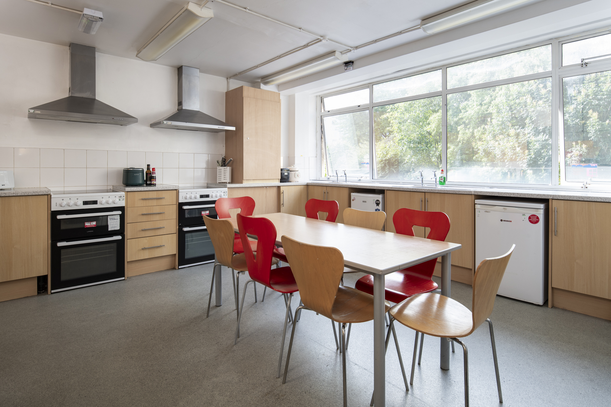 Shared kitchen area showing two refrigerators, two cookers and hobs, storage cupboards and a long table with eight chairs