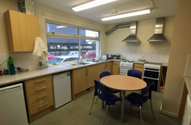 Shared kitchen area showing several refrigerators, two cookers and hobs, two sinks, storage cupboards and a table with four chairs