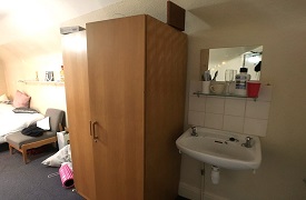 A bedroom with a small sink and wall mounted mirror. Next to the sink is a wardrobe, an armchair, and a bed.