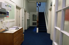 A blue carpeted hallway leading to a staircase at the far end. There is a chest of drawers and noticeboard to one side. 