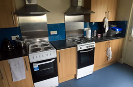 A kitchen with 2 ovens, 1 electric hob, 1 gas hob, and cupboard storage. On the countertop there are 2 kettles and a rice cooker.
