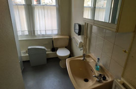 A bathroom with a sink, wall mounted mirrored cabinet and toilet. There is a large window at the far end of the room.