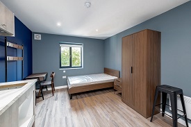 A studio room in The Skyline with a double bed in the right corner, a small bed side table to the right of the bed, a large wardrobe and a stool. To the left of the room there is a desk and chair against the wall.