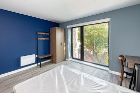 A studio room in The Skyline with large glass windows. A wardbrobe and storage area is in the right hand corner. A double bed is in the middle of the room and a desk with a chair is on the left hand side.