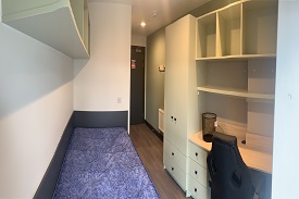 A bedroom with a single bed alongside the wall with shelving above. On the opposite wall there is a wardrobe, desk, office chair, and shelving.