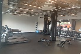 A gym with a full width mirror along the far wall. There is a rack with free weights by the mirror. On the opposite side of the room there are treadmills and a rowing machine. In the middle of the room there are some weight benches.