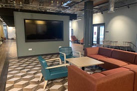 A large social area with a corner sofa and chairs around a coffee table. There is a large TV mounted in a wall in the middle of the room. There are chairs and tables placed alongside the length of the room by the wall.