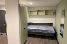 A bedroom with a double bed by the wall at the far end of the room. Above the bed there is some wall mounted storage. Next to the bed there is a wardrobe. Opposite the bed there is a wall mounted mirror.