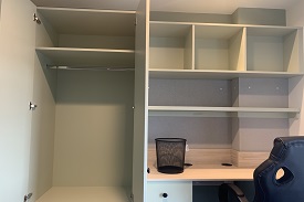 A study area alongside a wall in a bedroom. Inside the wardrobe there is a clothes rail. By the desk there is an office chair and shelving.