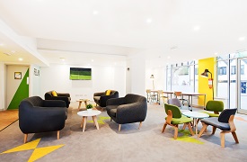 Social space, showing four sofas and two coffee tables, four chairs arranged around another coffee table, and three desks and chairs in the background. On the wall is mounted a screen