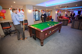 Social space, showing two students having a game of pool. The room is decorated with red bunting and balloons. In the background are several sofas and armchairs occupied by students