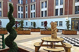A view of Unite House courtyard, surfaced in hard stone tiles and surrounded by the Unite House building itself. The courtyard has a number of circular benches with seating for up to eight people on each bench. The courtyard is decorated with wooden planters containing small tress and plants, and there are also three games - giant jenga, giant connect four, and giant chess