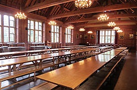 A large dining room containing three long rows of wooden tables with wooden bench style seating. The room has a wooden vaulted ceiling with ten lit chandeliers hanging from the beams. At the far end of the room six portraits hang from the wood panelled walls