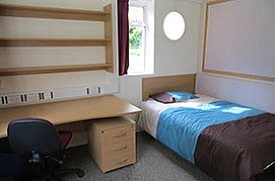 A room with a single bed, desk, chair, under desk storage, and shelves above the desk