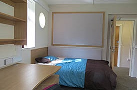 A room with a single bed, desk with chair, under desk storage and shelves above the desk, and a noteboard on the wall next to the bed