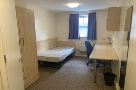 Small bedroom containing single bed, desk, chair, bin and fan, shelves above the desk, and cupboard. There is a radiator on the wall