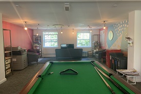 A room containing a pool table, with a sofa, three armchairs and a television at the far end. Along the walls are shelves, recycling bins and a photocopier
