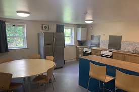 View of shared kitchen, containing a large refrigerator, two ovens/hobs, a sink, boiler, storage units, round table with five chairs, and two high chairs on an island unit