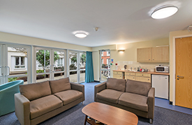 A common room with two sofas facing a coffee table. There is a kitchenette, and some double doors that open out on to the courtyard of Woodland Court.