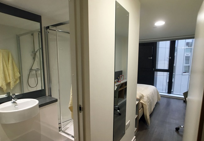 A room with a door leading into an en suite bathroom with a basin, mirror and shower cubicle. There is a mirror in the main room next to the bathroom door and a bed beyond that.