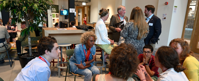 A group of six people sat down chatting. A group of four are also having a conversation in the background