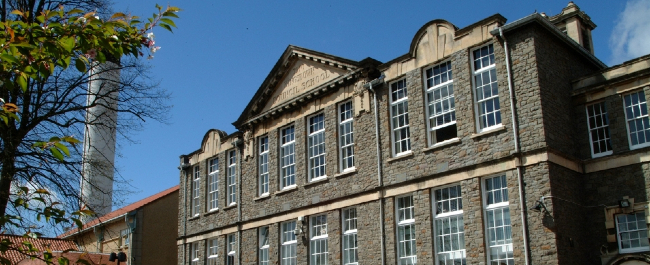 A landscape image of the School of Anatomy building under a clear blue sky. There is greenery in the foreground and a large industrial chimney in the background.