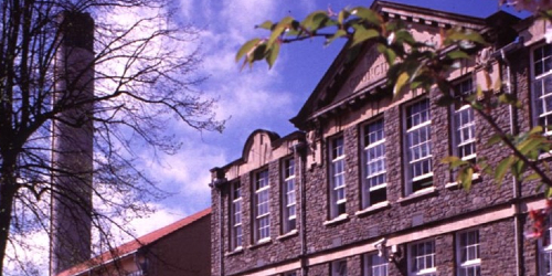 A landscape image of the School of Anatomy building under a clear blue sky. There is greenery in the foreground and a large industrial chimney in the background. Select to go to the School of Anatomy 'Virtual tour' page.