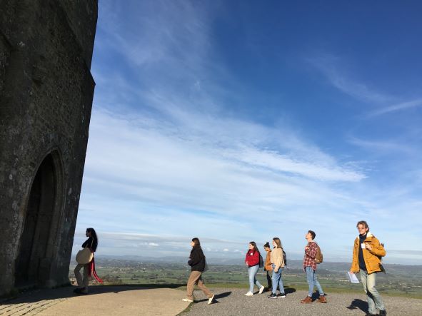 A group of students looking at a stone tower with view across fields, as part of their fieldtrip to Glastonbury